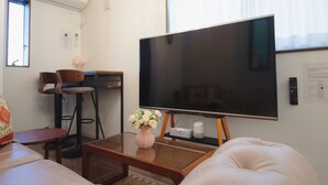 A living room with a large flat-screen TV, a sofa, a coffee table with a vase of flowers, a chair, and a small table with a remote control.