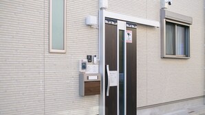 A building with a light-colored brick facade, a door with a security camera, and a window with a curtain.