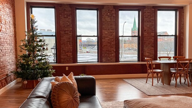 A cozy living room with a leather sofa, a small Christmas tree, and a dining area with wooden chairs and a table. - Luxurious 2 BR Loft  (Utica)