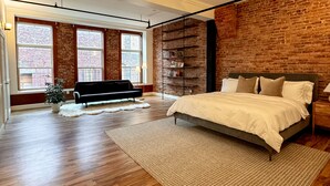 A bedroom with a large bed, a black sofa, a brick wall, and a window with a view of a brick building. - Luxurious 2 BR Loft  (Utica)