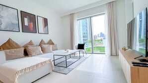 Una sala de estar moderna con un sofá, una mesa de centro y un televisor. Hay ventanas grandes con cortinas, y vista a un paisaje urbano.