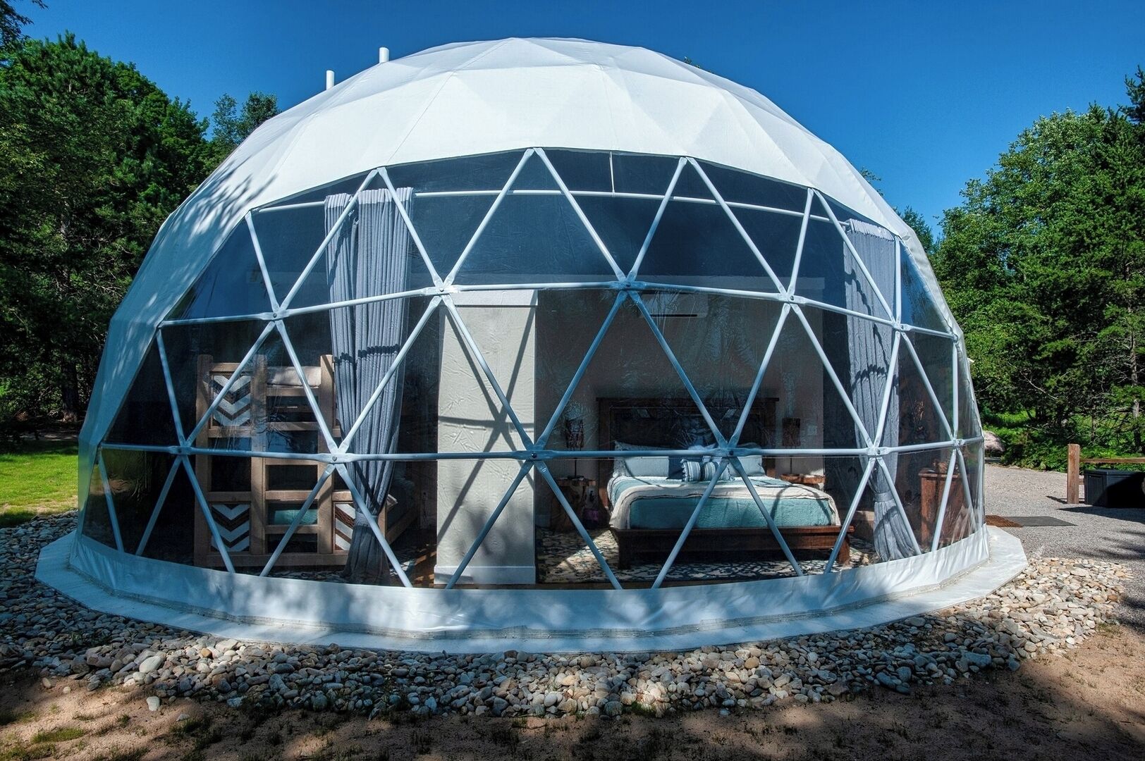 A geodesic dome structure with a central bed and seating area, surrounded by a gravel path and trees.