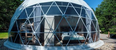 A geodesic dome structure with a central bed and seating area, surrounded by a gravel path and trees.