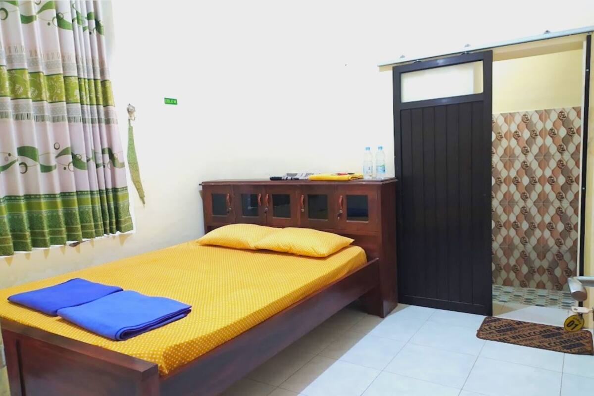 Un lit simple avec un matelas jaune et une couverture bleue, une armoire en bois, une petite table recouverte d’une nappe jaune et une porte coulissante agrémentée d’un rideau à motifs.