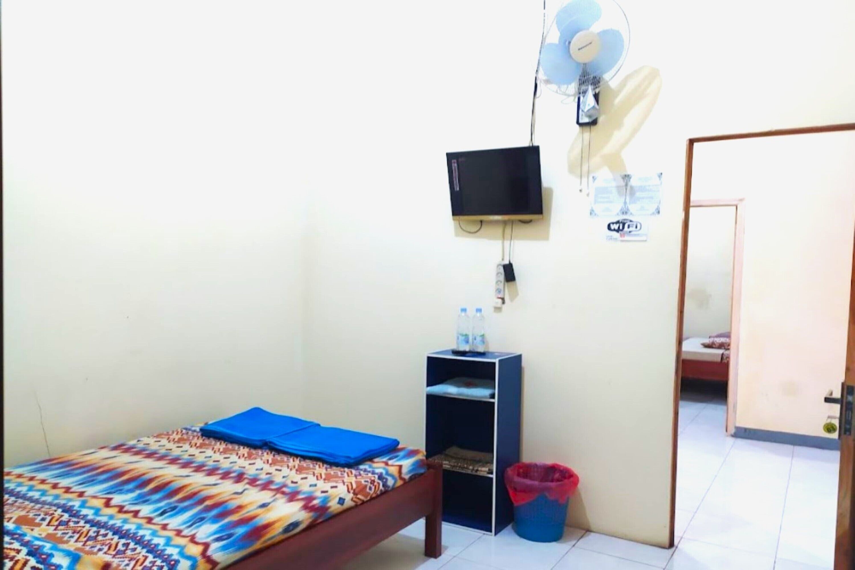 A small, simple room with a bed, a TV, a shelf with water bottles, and a trash bin.