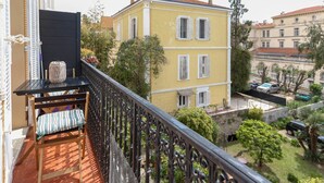 A balcony with a black table and a cushioned bench, overlooking a courtyard with a yellow building and greenery. - Modern apartment with 3 bedrooms, balcony, communal garden and A/C- Benakey (Cannes)