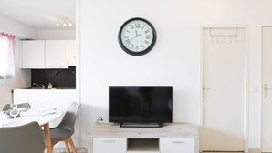 Una sala de estar moderna con un televisor de pantalla plana sobre un mueble de madera, un comedor con una mesa y sillas blancas, y una cocina al fondo.