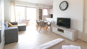 Una sala de estar moderna y bien iluminada, con un sofá, televisor, comedor con mesa y sillas, y una zona de cocina con armarios.