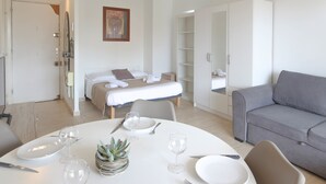 Una habitación moderna y bien iluminada con una cama impecable, un sofá gris y una zona de comedor con platos, vasos y cubiertos.