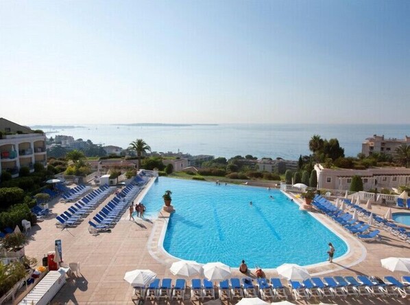 Une grande piscine extérieure, avec des chaises longues bleues et des parasols, entourée de bâtiments et offrant une vue sur la mer.