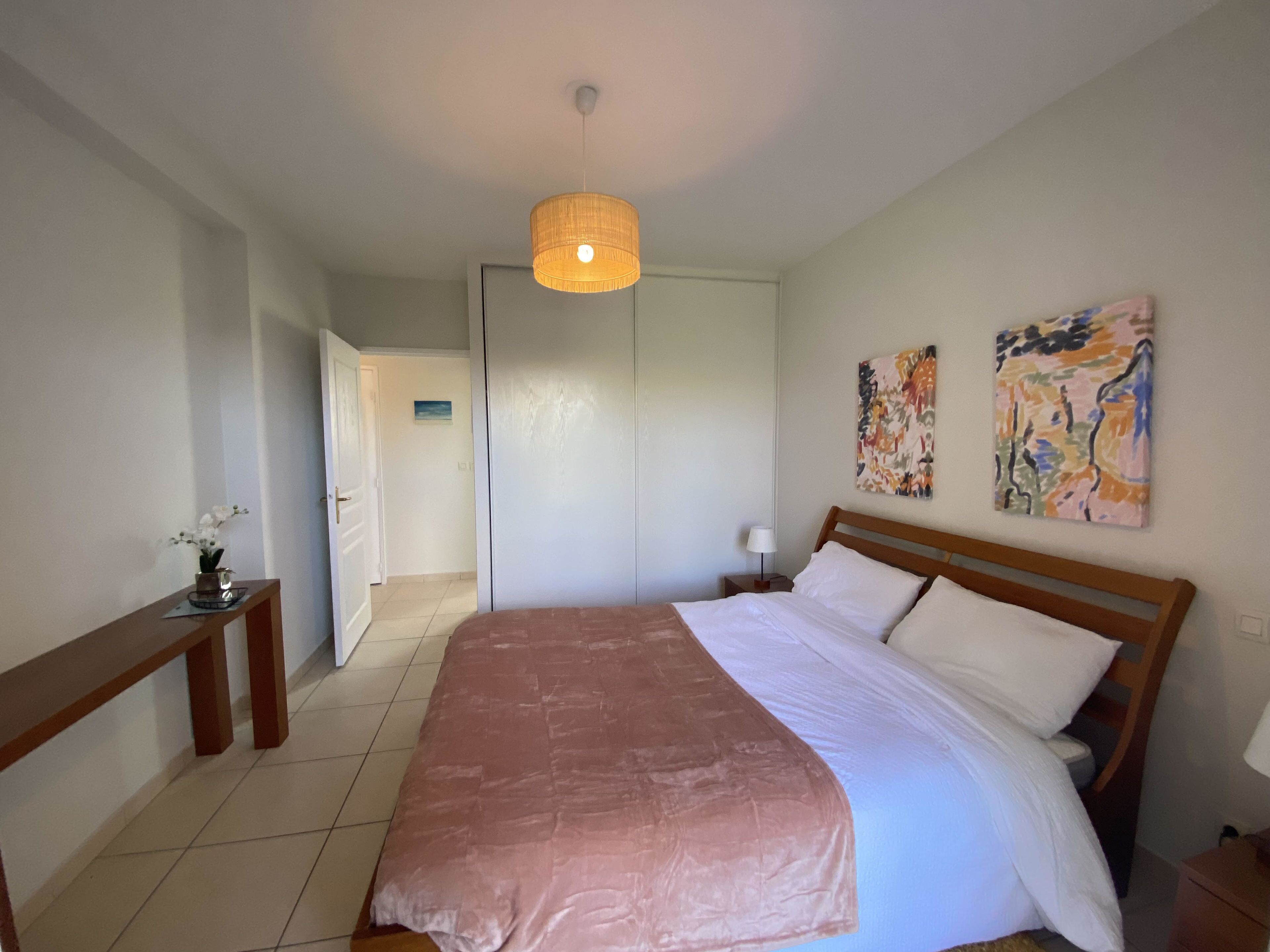 A bedroom with a large bed, a bedside table, a hanging light fixture, and two wall art pieces.