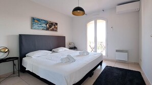 Ein ordentlich bezogenes Bett mit weißen Leinen und einem aufgerollten Handtuch, ein schwarzer Kopfteil, ein kleiner Nachttisch mit Lampe, ein Gemälde an der Wand und ein Blick ins Freie durch eine Glastür.