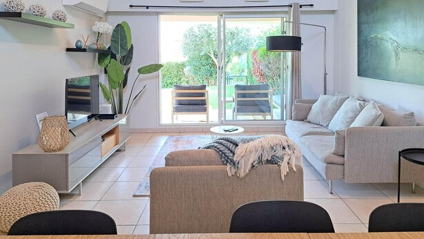 Ein modernes Wohnzimmer mit einer Couch, einem Couchtisch und einem Fernseher auf einem Schrank. Eine Schiebetür aus Glas führt in einen Außenbereich mit einem Stuhl und einem Tisch.