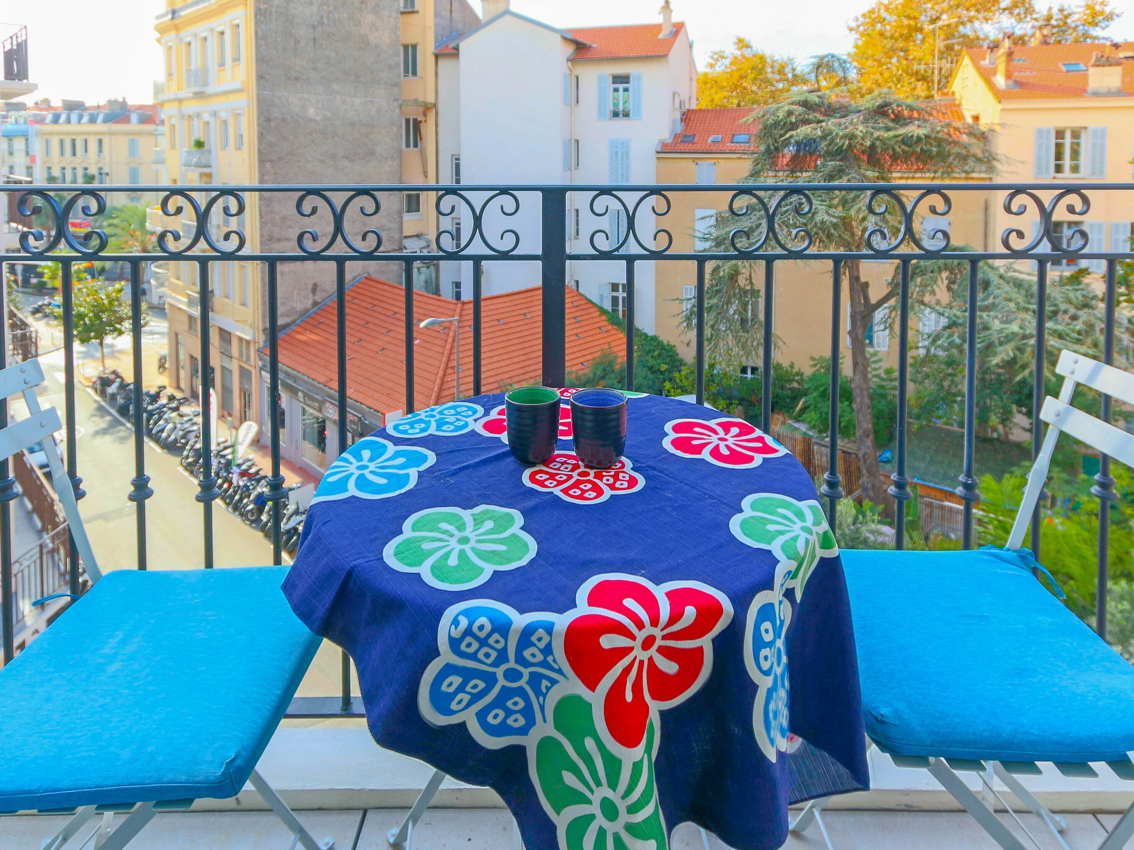 A balcony with a table covered by a floral tablecloth, two blue cushioned chairs, and a metal railing.