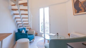 Un salon moderne avec une table basse en verre, un canapé bleu, un fauteuil blanc, un escalier avec des marches en bois et un grand tableau accroché au mur.