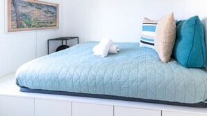 Un lit bien fait, recouvert d’une courtepointe bleue matelassée, avec une serviette blanche et deux oreillers (un à rayures et l’autre uni, de couleur turquoise). Un tableau encadré est visible sur le mur, à gauche.