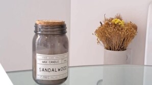 Un bocal étiqueté « bougie au bois de santal » est posé sur une table en verre, à côté d’un vase blanc contenant des fleurs séchées.