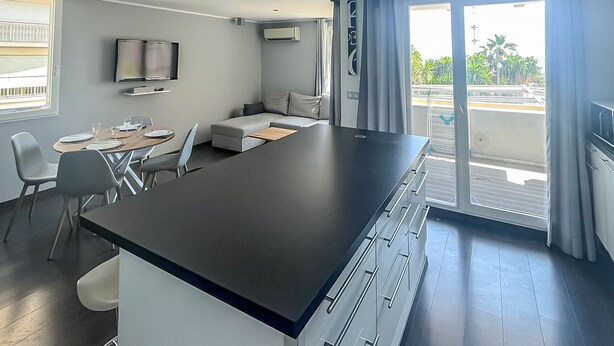 Une cuisine moderne dotée d’un îlot central, d’un coin repas avec des chaises, d’un canapé et d’un téléviseur fixé au mur.