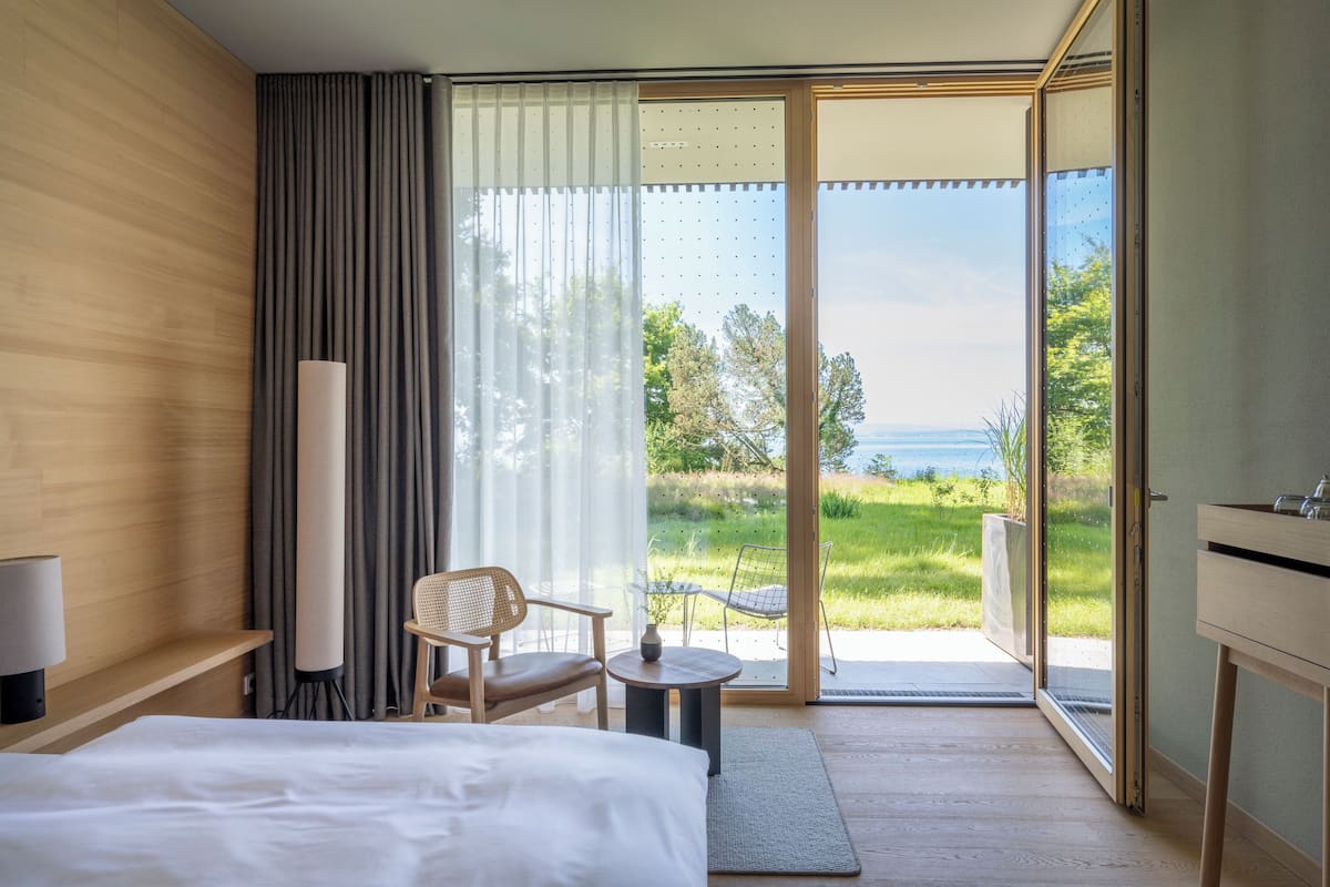 A hotel room with a bed, a chair, a small table, and a view of the outdoors.