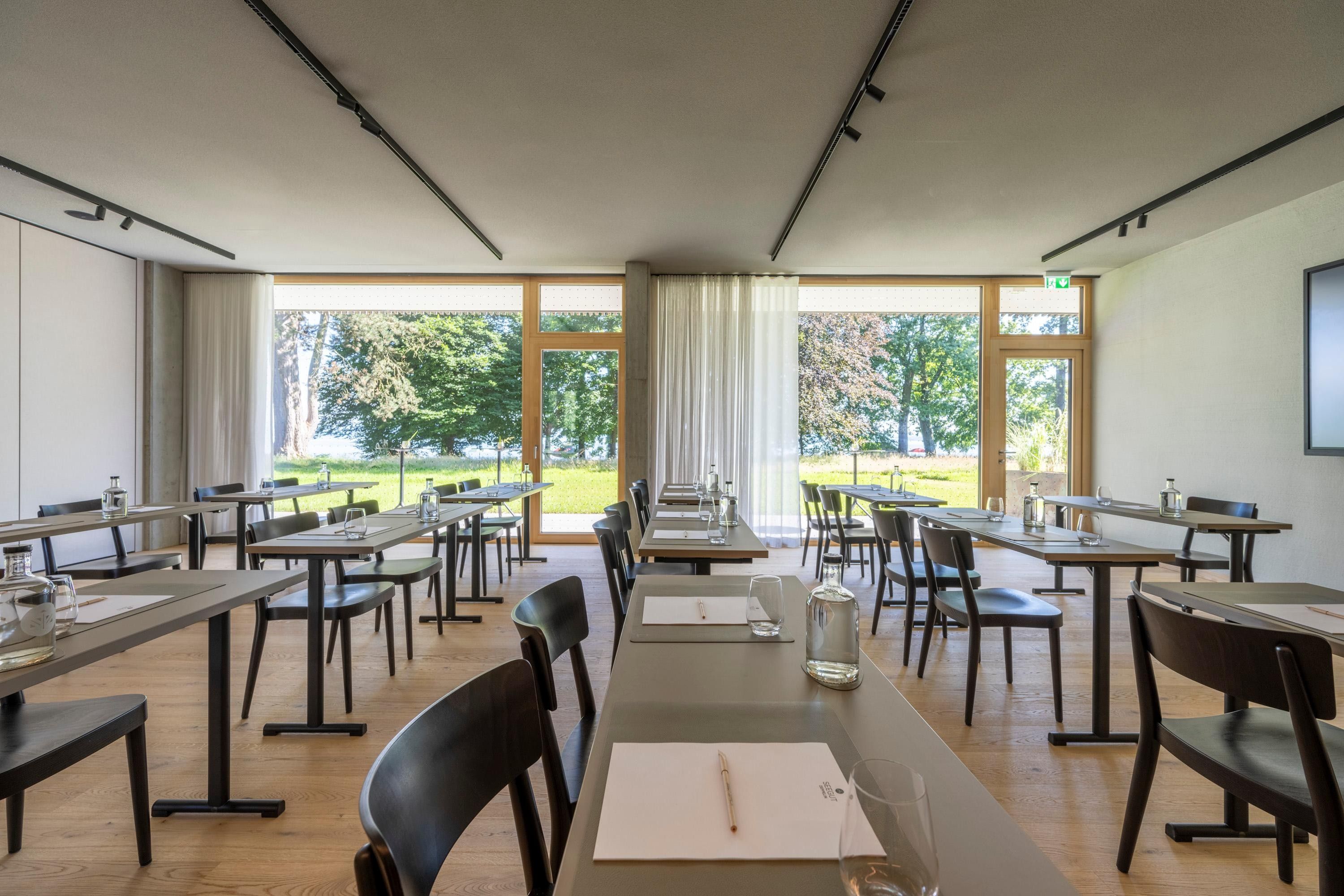 A conference room with multiple tables and chairs, large windows with curtains, and a view of greenery outside.