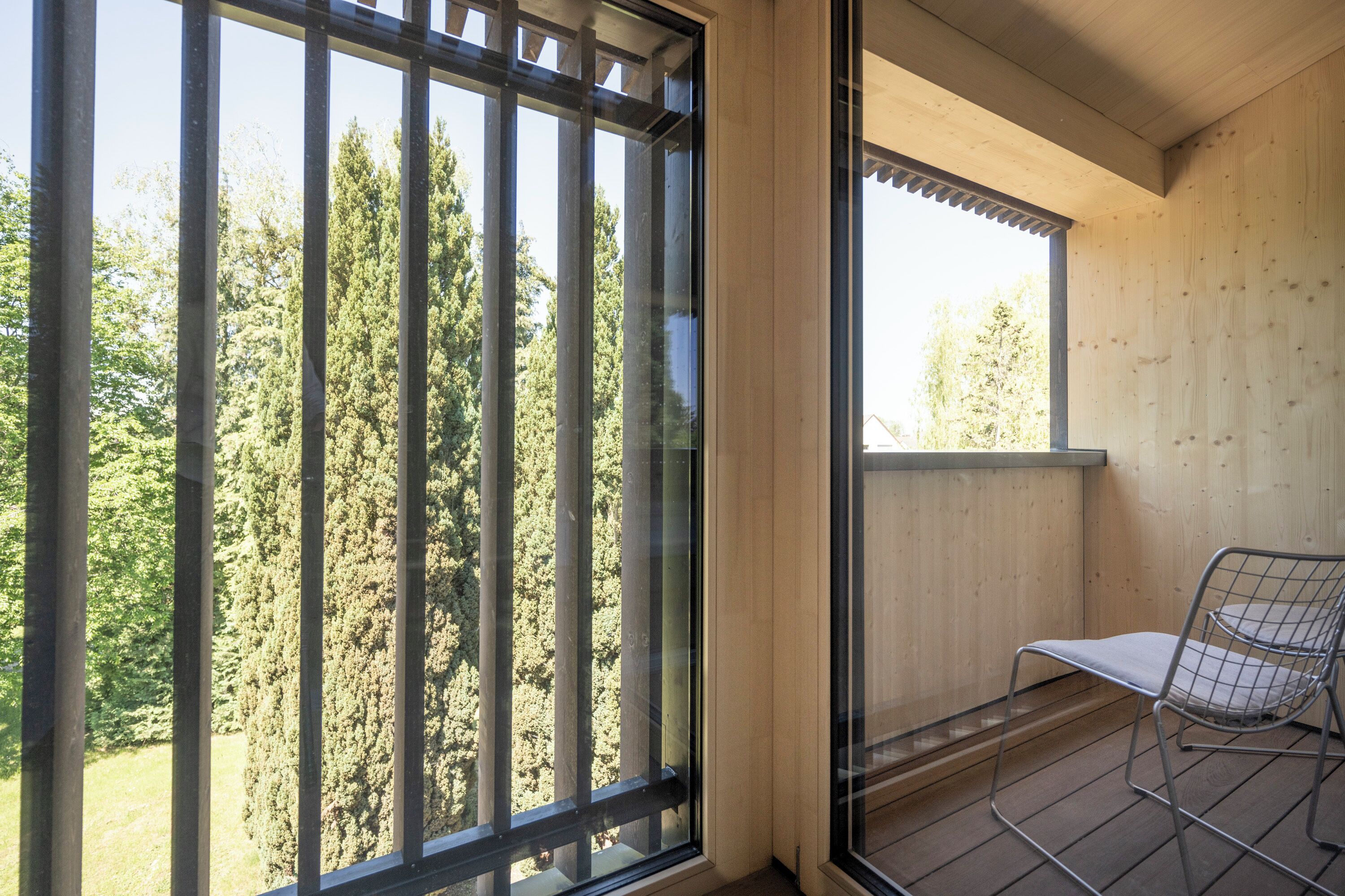 Een balkon met een metalen stoel, grote ramen en uitzicht op bomen.