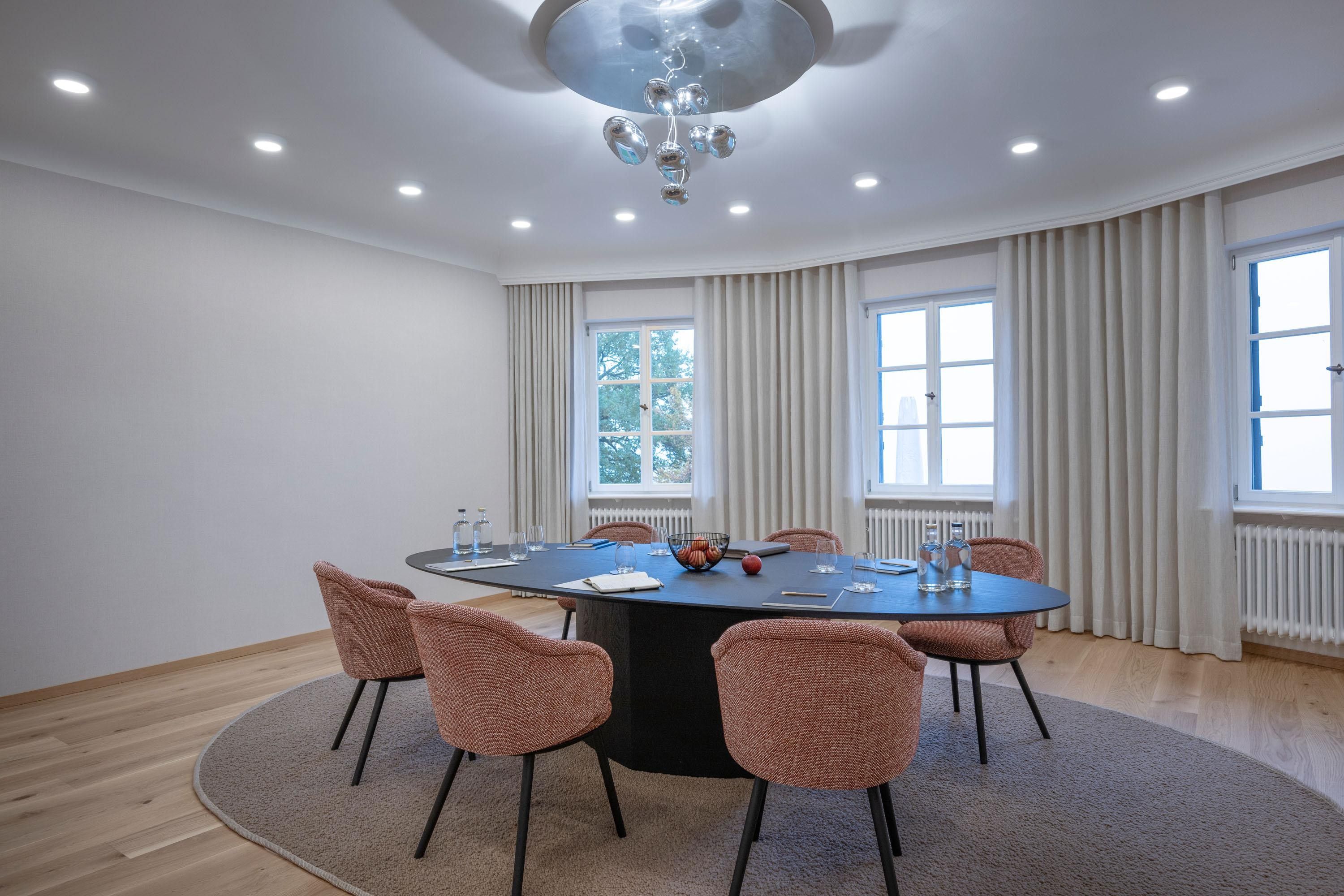 A conference room with a round table, six chairs, a chandelier, and large windows with curtains.