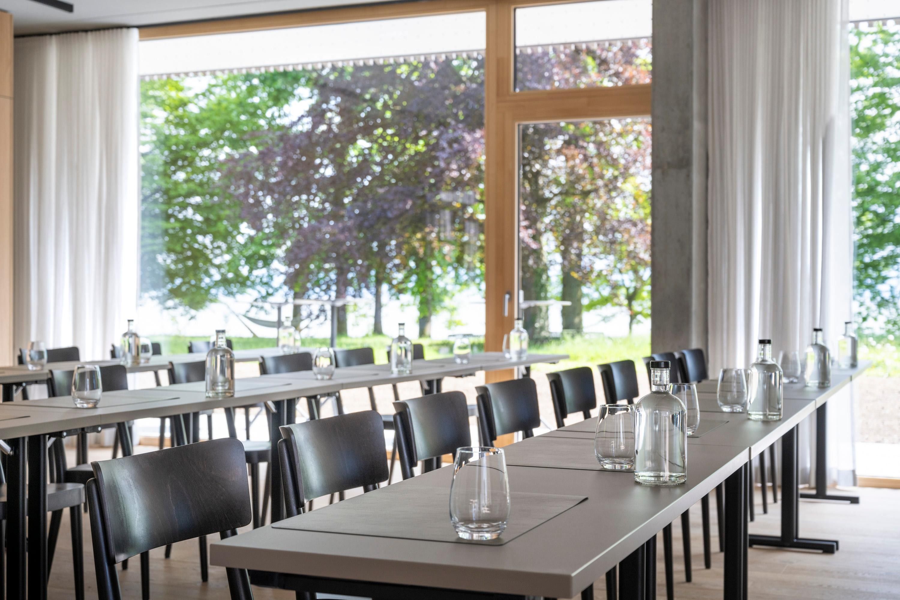 A long dining table with chairs, clear glassware, and bottles, set in a room with large windows and curtains.