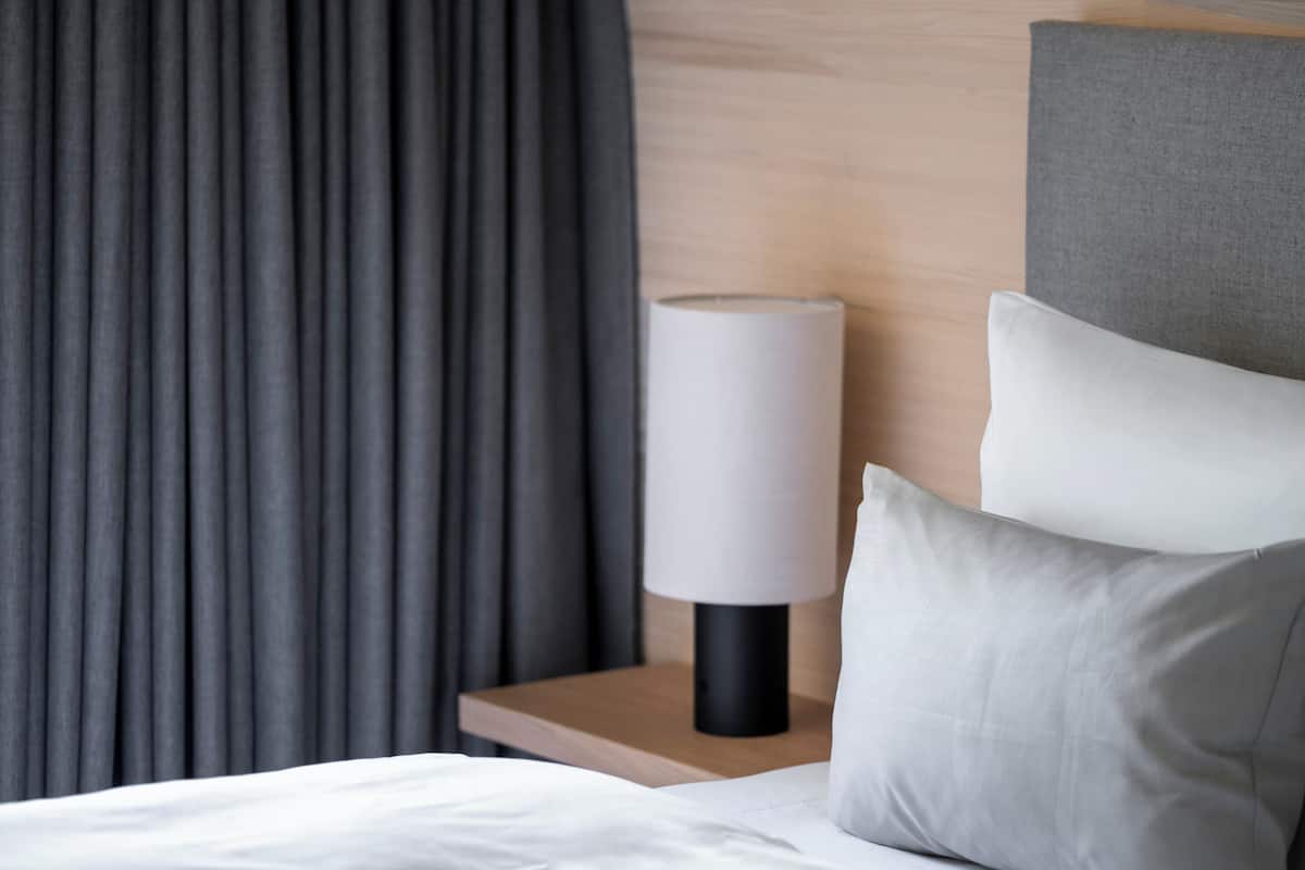 A neatly made bed with white linens and a grey pillow, a bedside table with a lamp, and grey curtains.