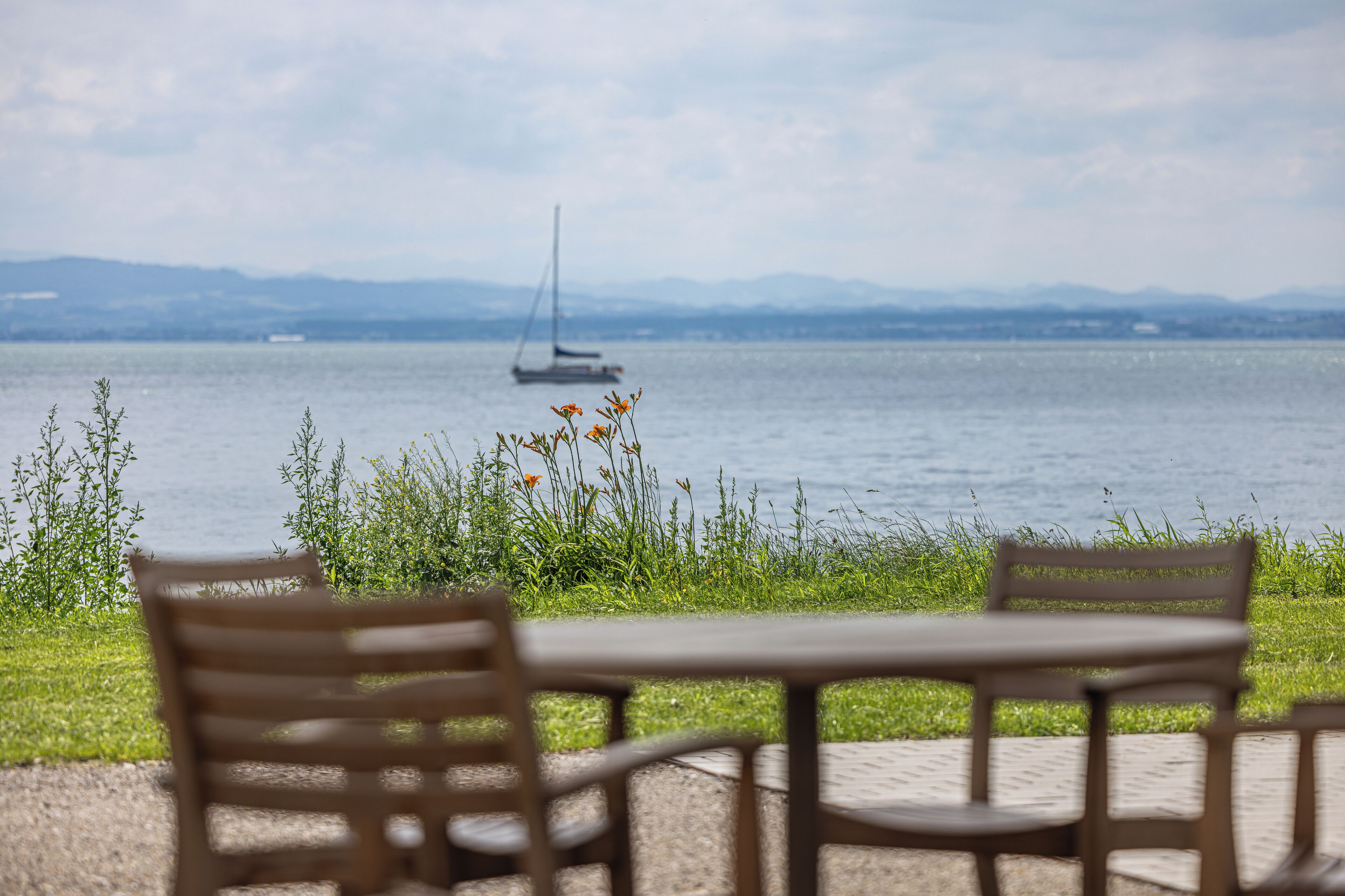 Una mesa y sillas junto a un lago, con un velero a lo lejos.