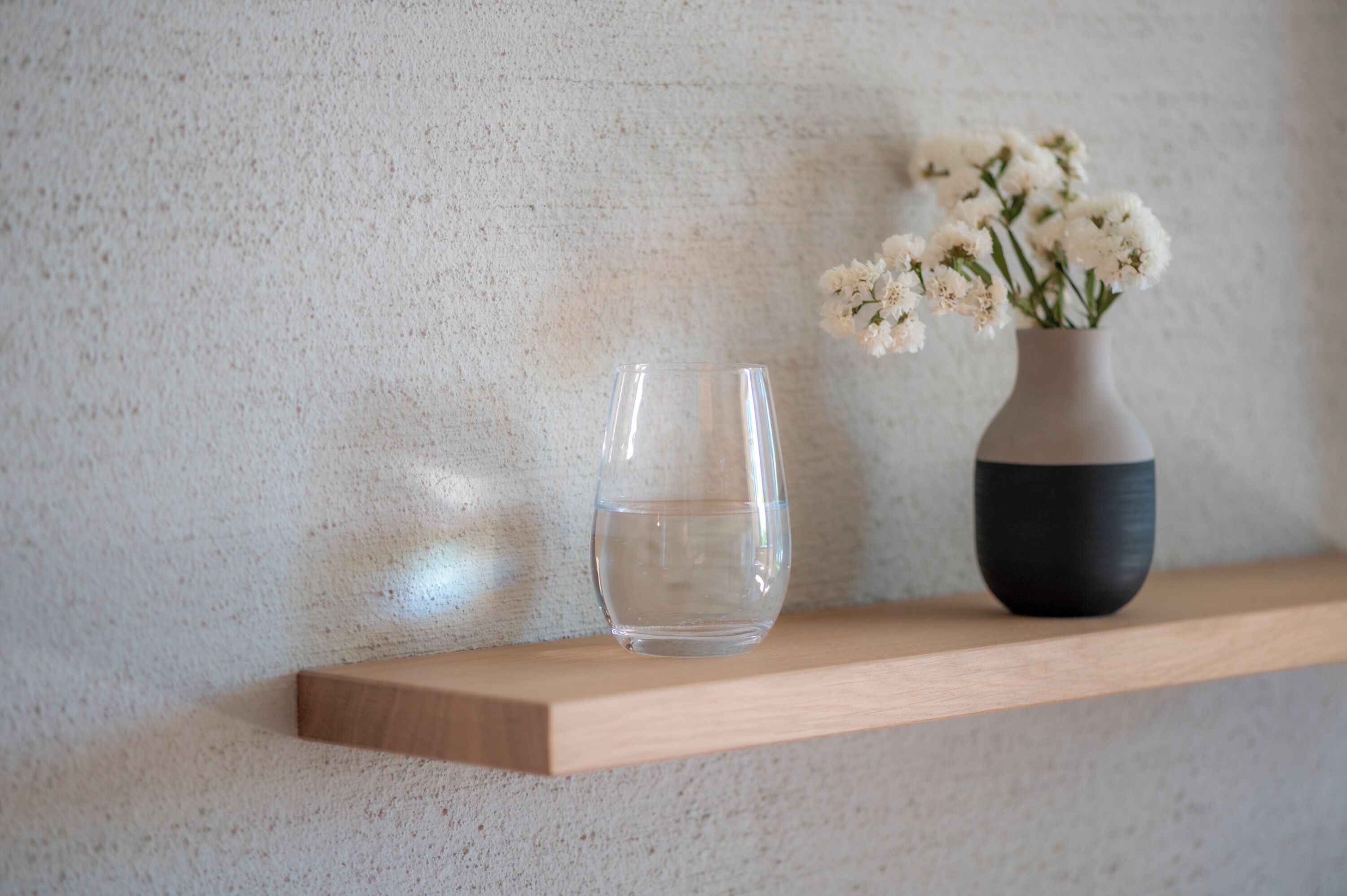 Un verre transparent et un vase rempli de fleurs sont posés sur une étagère en bois, devant un mur à la texture particulière.