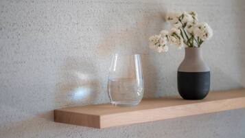 Un verre transparent et un vase rempli de fleurs sont posés sur une étagère en bois, devant un mur à la texture particulière.