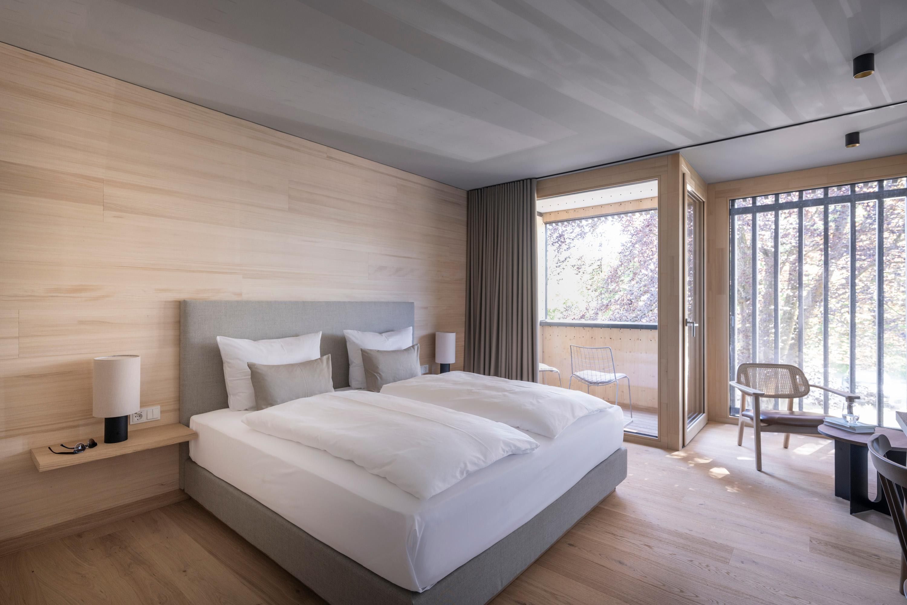 A modern bedroom with a large bed, bedside lamps, and a view of the outdoors through a sliding glass door.