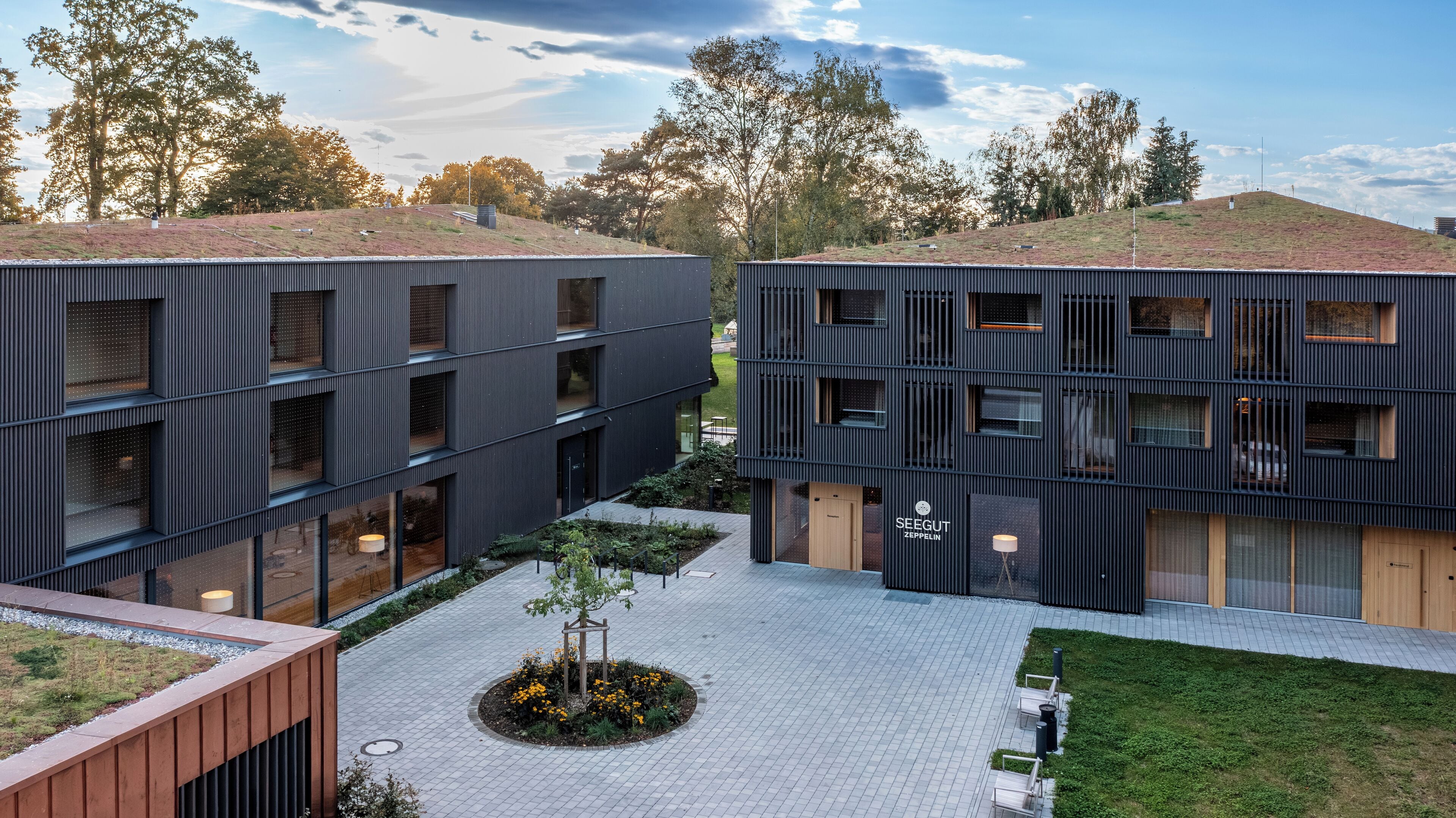 Un bâtiment moderne à plusieurs étages, doté de grandes fenêtres et d’une cour intérieure agrémentée d’un petit arbre et d’un parterre de fleurs.