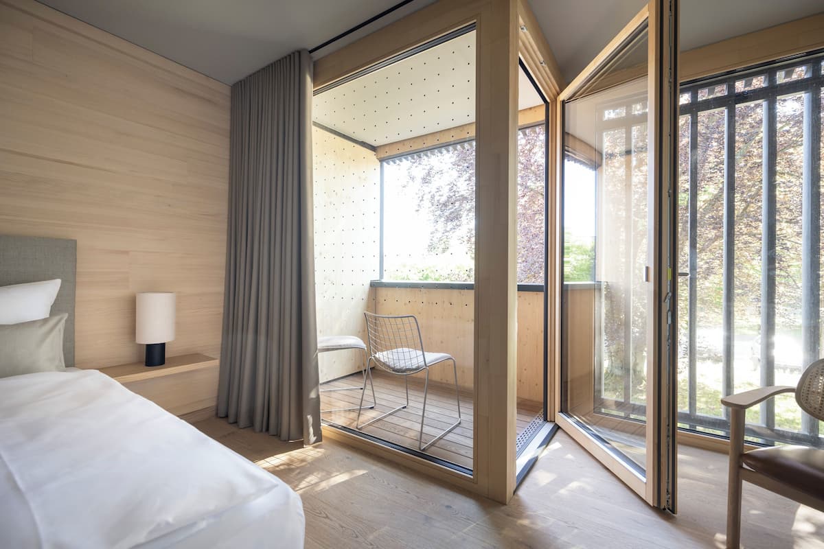 A modern hotel room with a large bed, a bedside table with a lamp, a chair, and a sliding glass door leading to an outdoor balcony.