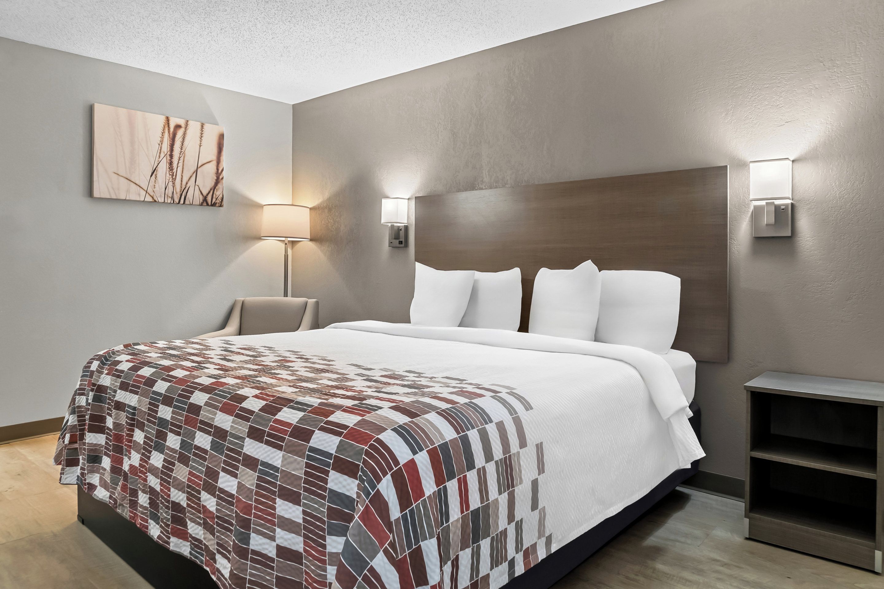 A modern hotel room with a large bed, a headboard, two wall-mounted lamps, a bedside table, and a small nightstand.