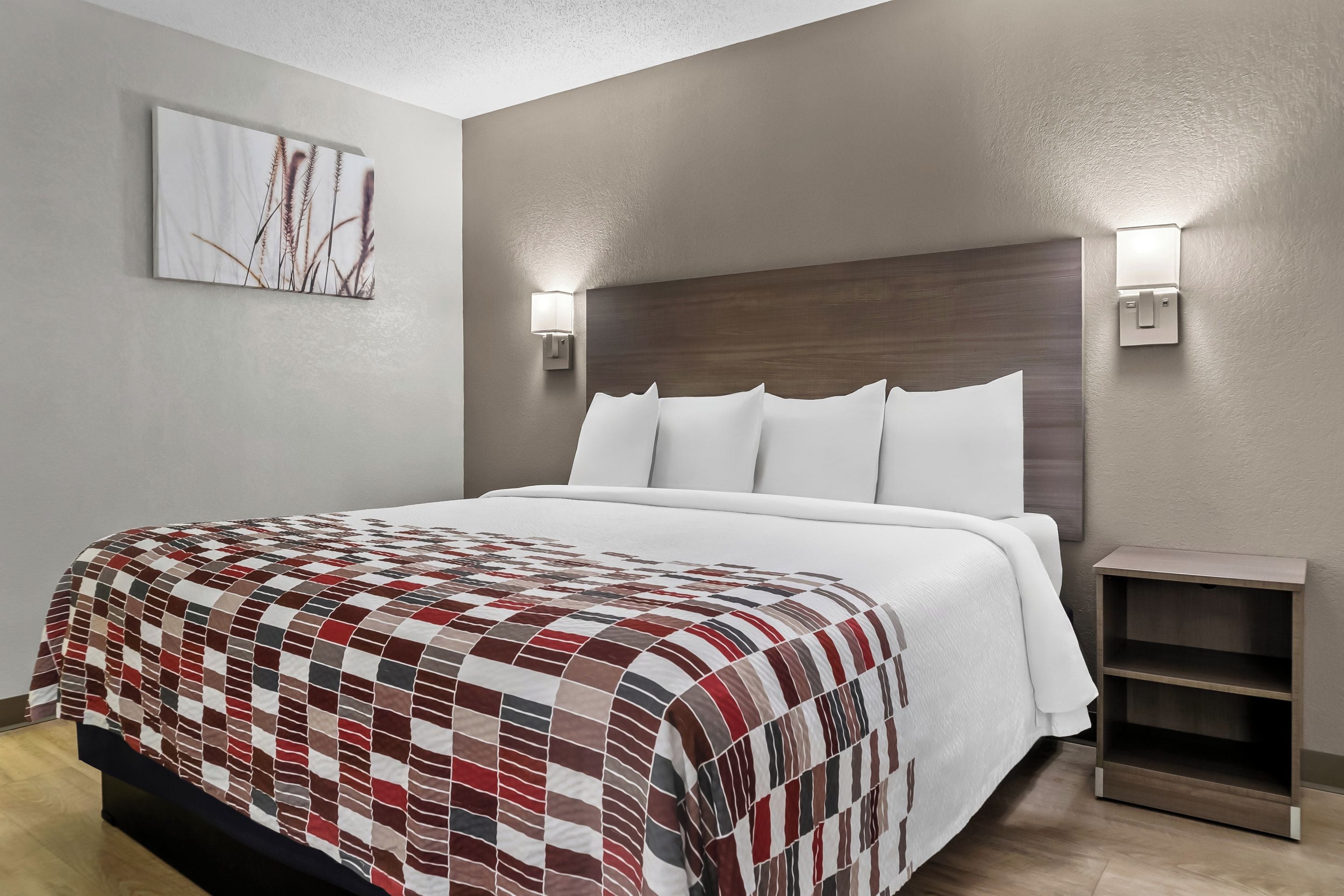 A hotel room with a large bed, a headboard, two wall-mounted light fixtures, a small wooden nightstand, and a wall-mounted artwork.