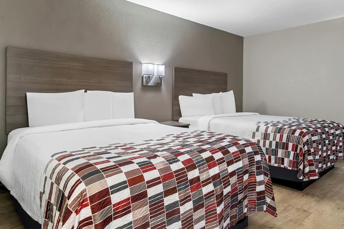 A hotel room with two beds, each with white bedding and a red and white checkered blanket on one bed. There is a wall-mounted light fixture above the beds.