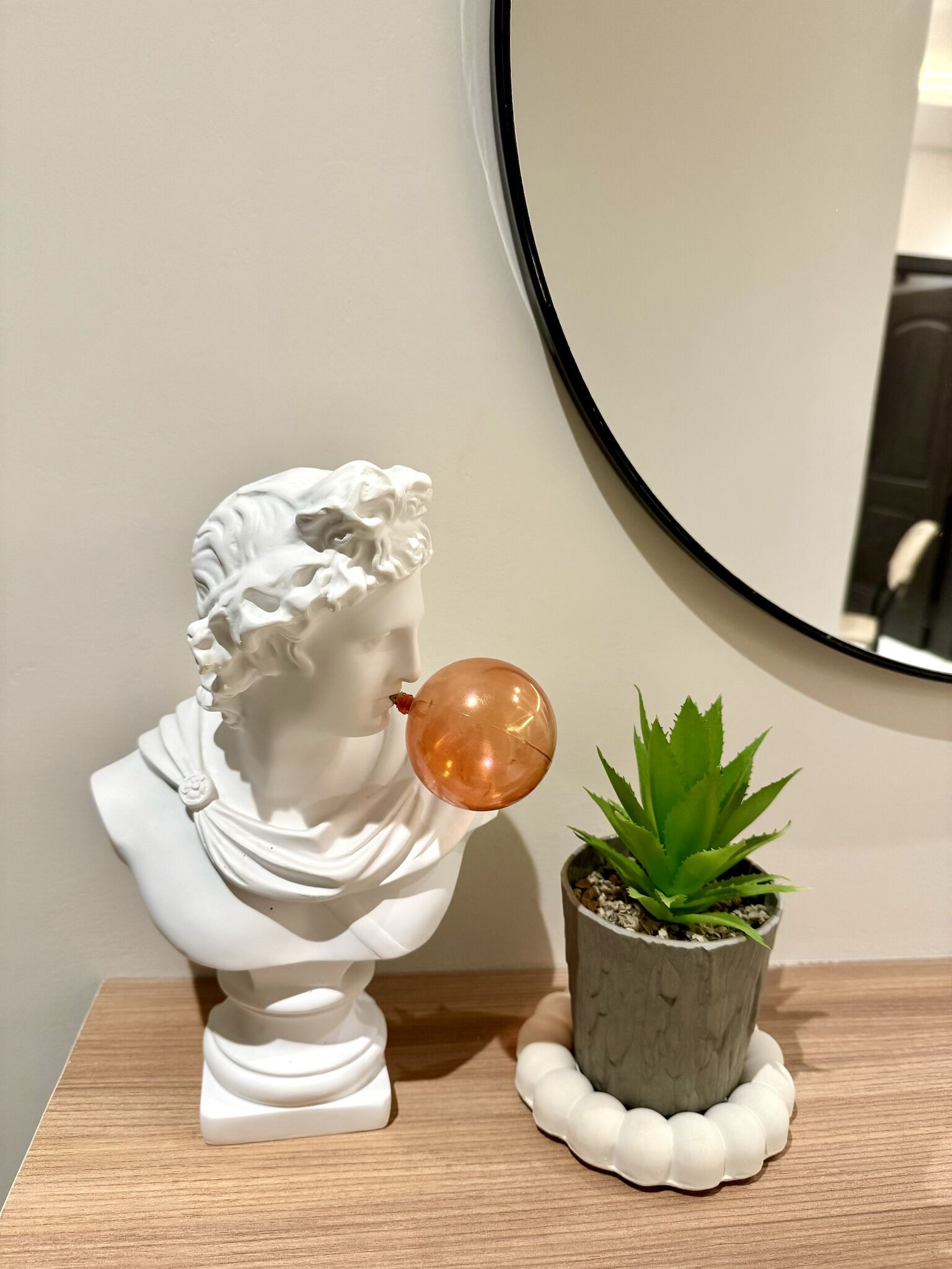 Escultura bust blanca amb un globus, una petita planta en test i un mirall rodó sobre una superfície de fusta.