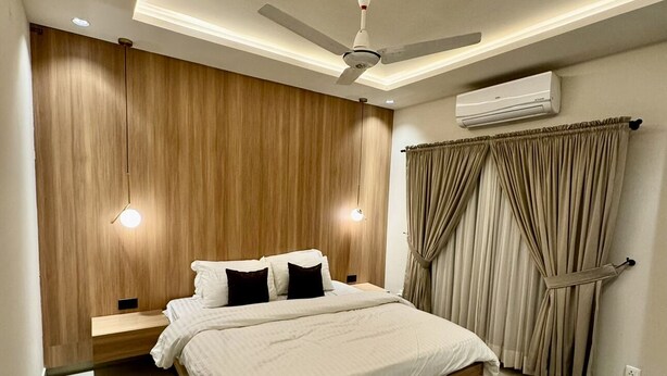 Quarto moderno com cabeceira de madeira, cama com roupa de cama branca, duas luminárias de cabeceira, ventilador de teto e cortinas bege.