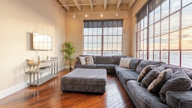 A spacious living room with a large sectional sofa, a small ottoman, and a console table with decorative items. - Luxurious Downtown Condo w/ Panoramic City Views and Free Secure Onsite Parking. (New Orleans)
