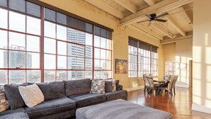 A spacious living room with a large gray sectional sofa, a matching ottoman, and a dining area with a wooden table and chairs. - Luxurious Downtown Condo w/ Panoramic City Views and Free Secure Onsite Parking. (New Orleans)