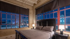 A bedroom with a large bed, two bedside tables, and a view of the city through large windows. - Luxurious Downtown Condo w/ Panoramic City Views and Free Secure Onsite Parking. (New Orleans)