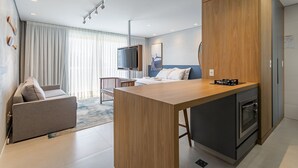 A modern hotel room with a kitchenette, a sofa, a TV, and a large window with curtains. - WI-FI 300MB | partial view of the SEA #CAMP09 (Florianópolis)