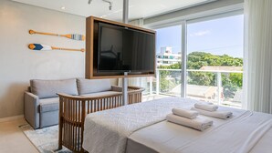 A modern hotel room with a large bed, a flat-screen TV mounted on the wall, a seating area with a sofa and armchair, and a view of the city through large windows. - WI-FI 300MB | partial view of the SEA #CAMP09 (Florianópolis)