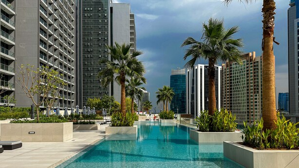 Un espace piscine moderne avec deux chaises longues blanches, entouré de palmiers et de grands immeubles.