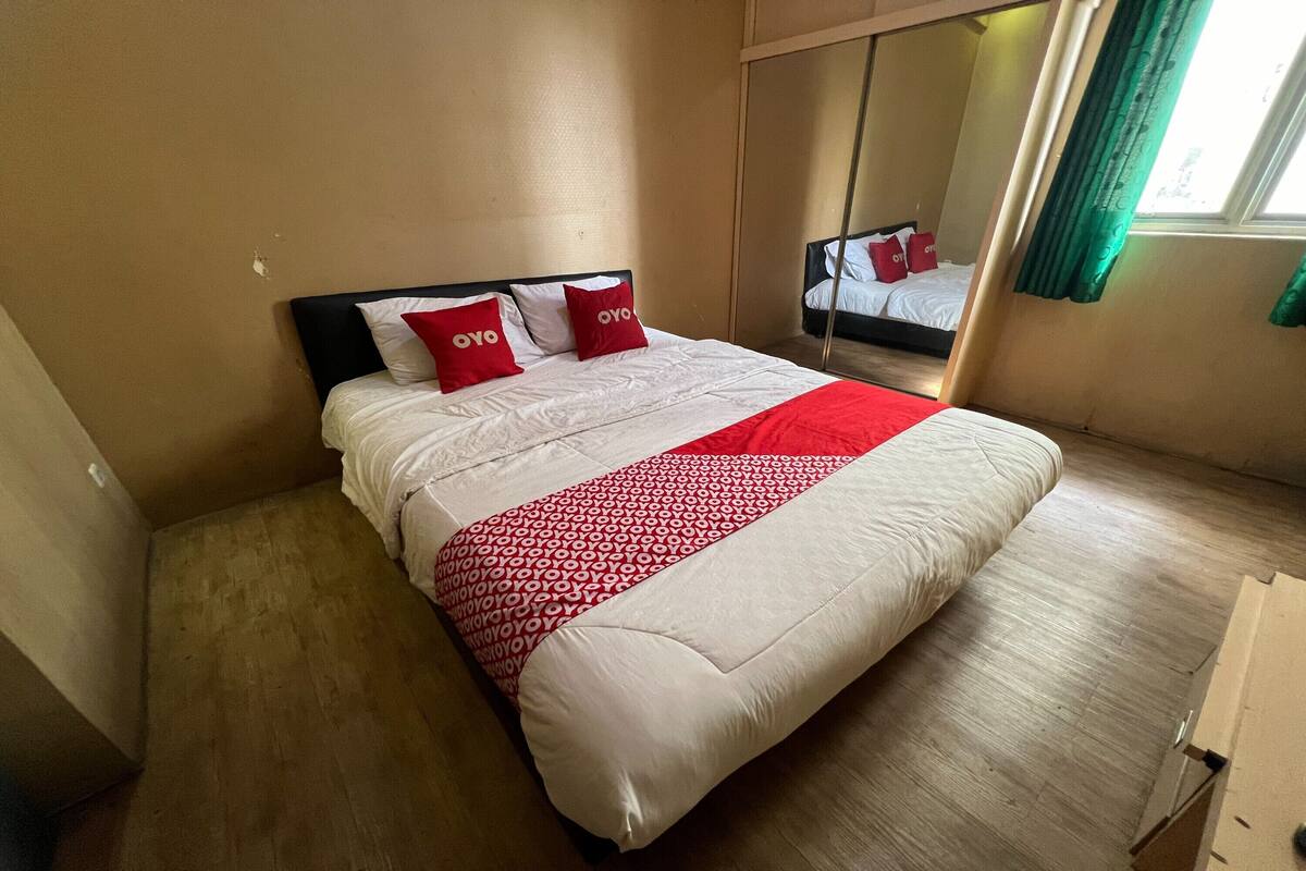 Una cama bien hecha con una colcha roja y blanca, dos almohadas rojas y un espejo grande en la pared.