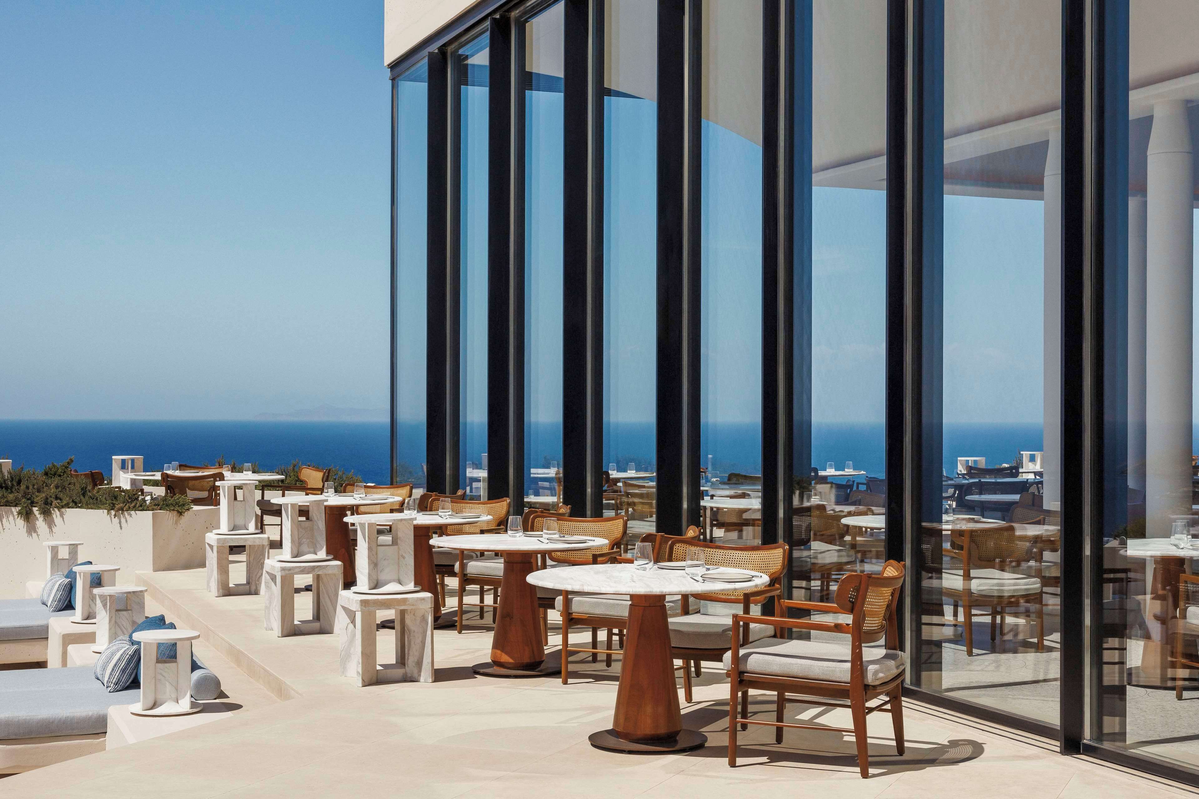 A rooftop terrace with round tables and chairs, large glass windows, and a view of the ocean.