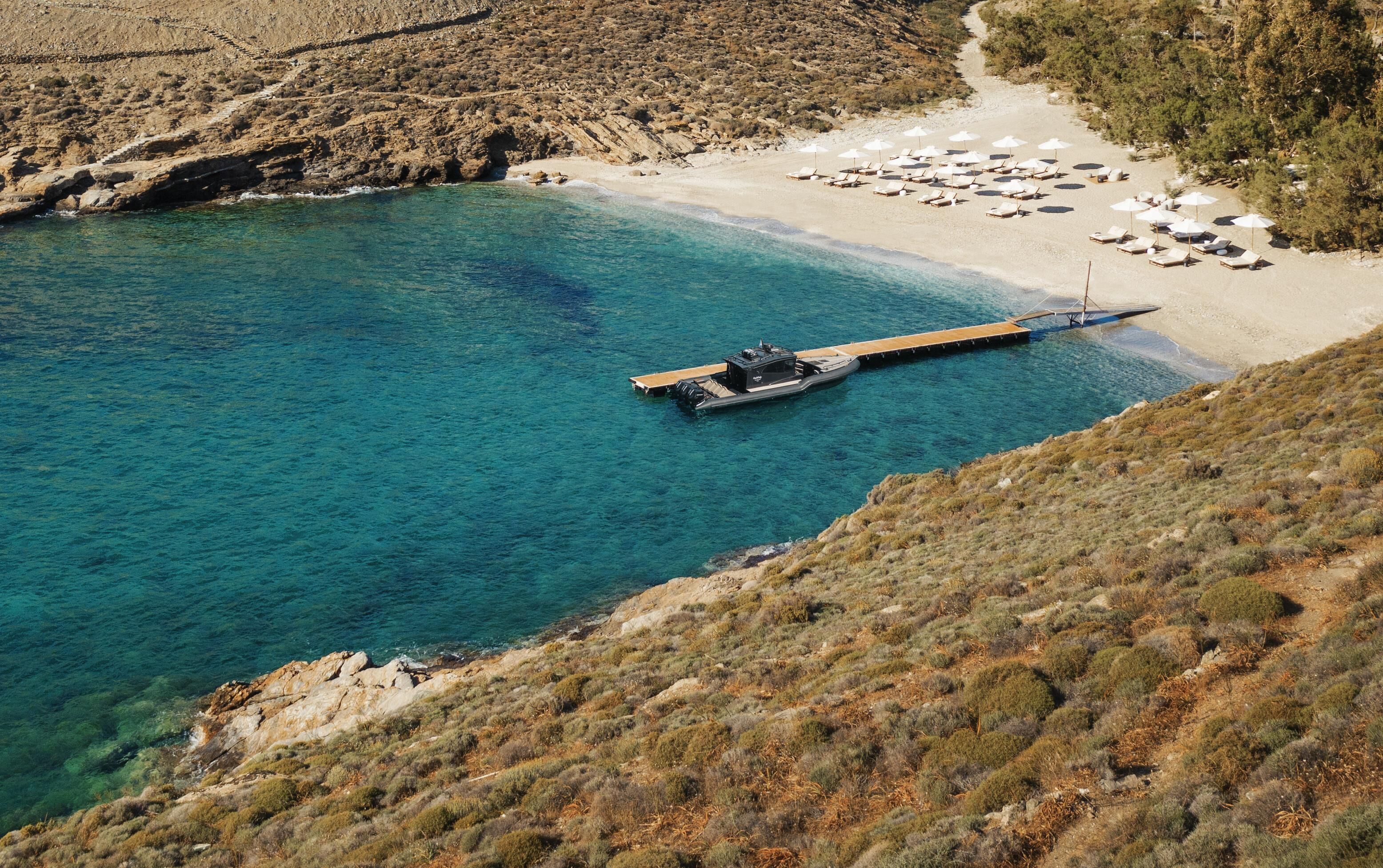 Vista aérea de una zona costera con un muelle que se adentra en el mar, rodeada por una playa con tumbonas y sombrillas.