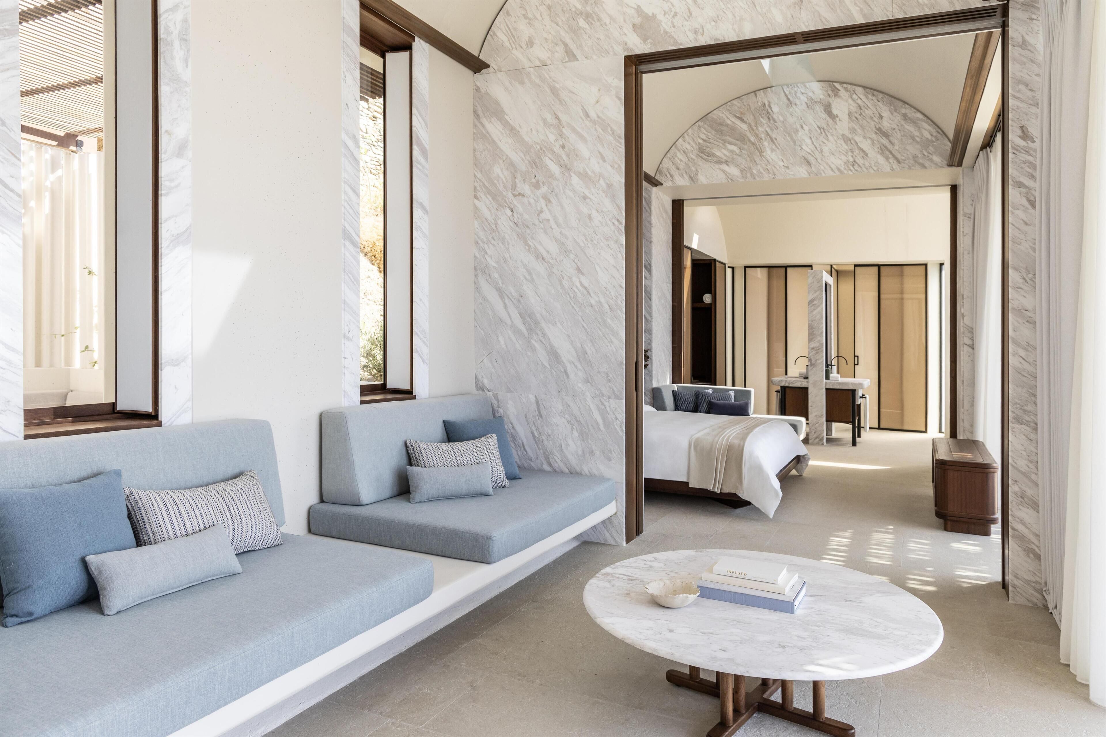 A modern hotel room with a marble floor, a sofa with blue cushions, a round marble coffee table, and a bed with a white headboard.