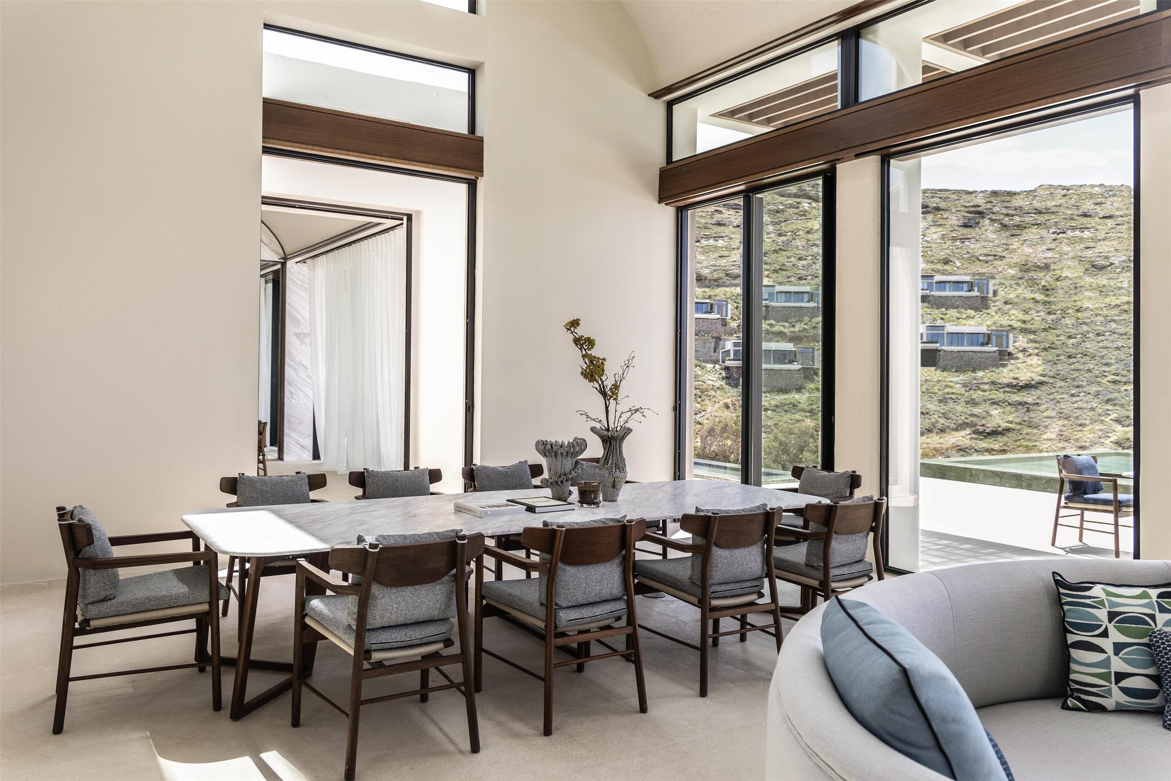 A modern dining area with a large table and chairs, large windows, and a view of a mountainous landscape.